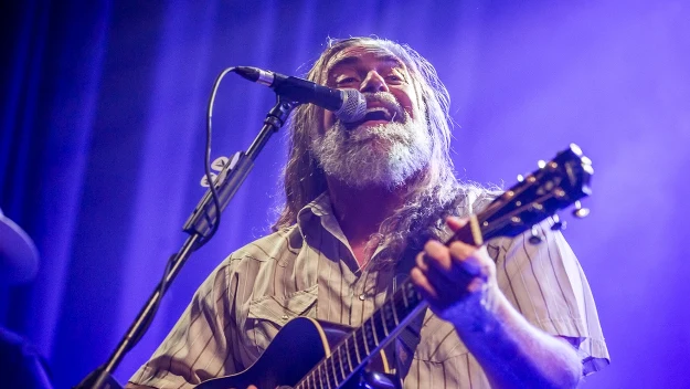 Starszy mężczyzna z długimi, siwymi włosami i brodą, śpiewający do mikrofonu podczas gry na gitarze akustycznej, oświetlony niebiesko-fioletowym światłem scenicznym.