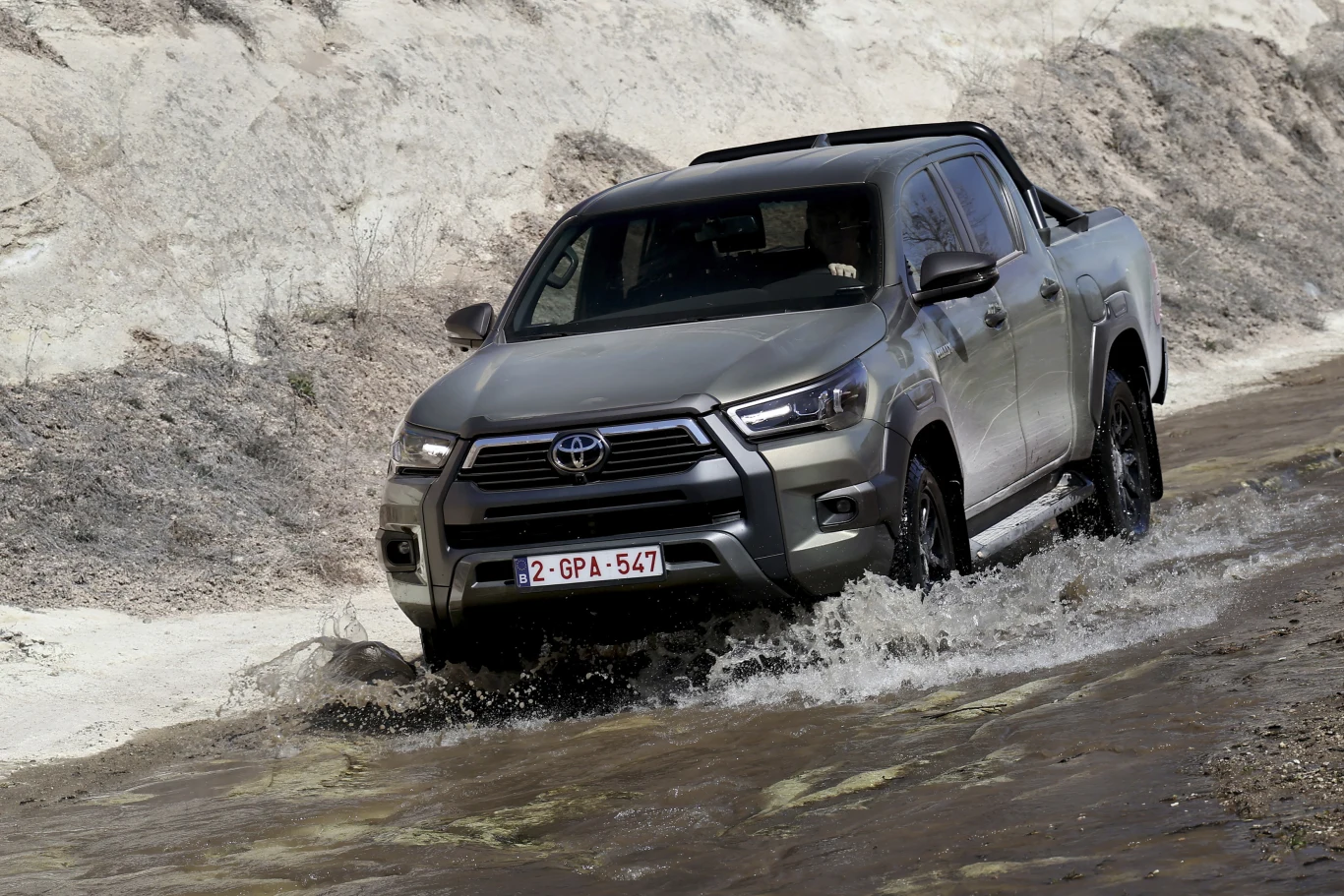 Toyota Hilux schodzącej generacji jest teraz dostępna z dużymi rabatami Srebrna Toyota Hilux pokonuje płytki strumień, unosząc wodę spod kół w terenie o jasnej, skalistej nawierzchni.