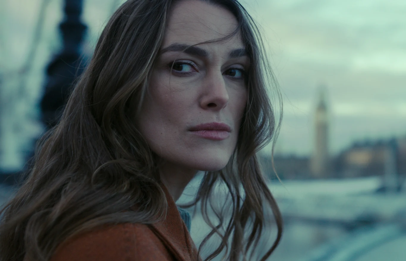 Keira Knightley w serialu "Black Doves" Keira Knightley jako Helen Webb w serialu 'Black Doves', patrzy przez ramię, w tle miasto i wieża zegarowa.