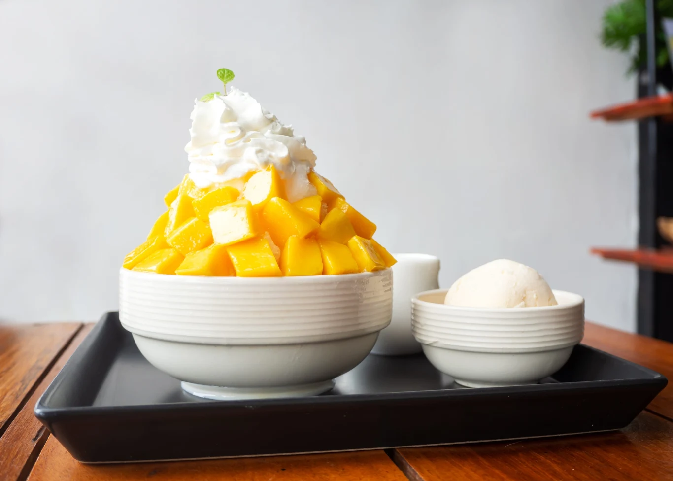 Bingsu można podać niemal z każdym dodatkiem: owocami, syropami, sosami, matchą czy ciastkami Deser z kawałkami świeżego mango, bitą śmietaną na wierzchu, podany w białej misce obok gałki lodów waniliowych, całość na czarnej tacy.