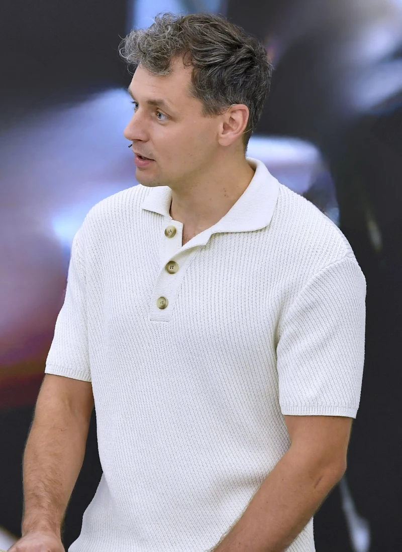 Łukasz Bożycki Mężczyzna w jasnym swetrze z krótkim rękawem i kołnierzykiem, stojący bokiem, z lekko uniesioną głową, na rozmytym tle.