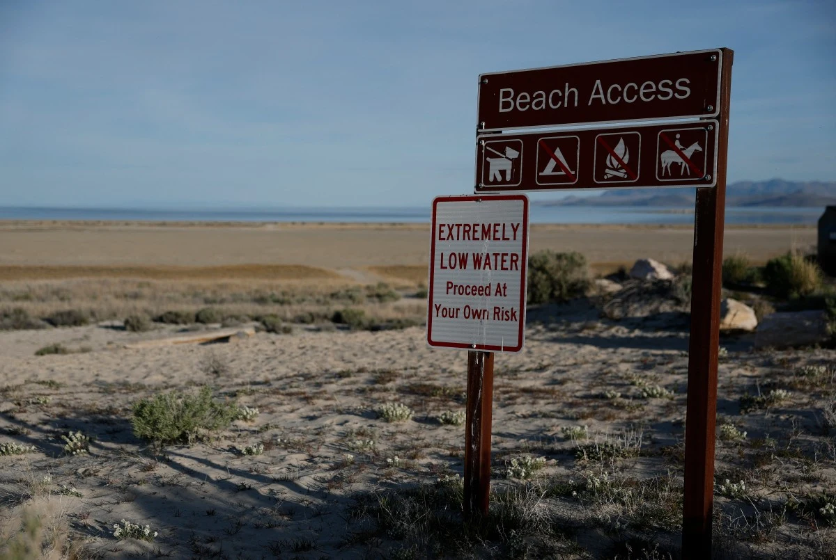 Tablica informująca o niskim stanie wód w jeziorze Brązowe tablice informacyjne z napisem 'Beach Access' oraz ostrzeżeniem o ekstremalnie niskim poziomie wody, ustawione na piaszczystym terenie prowadzącym w stronę szerokiego, suchego wybrzeża jeziora.
