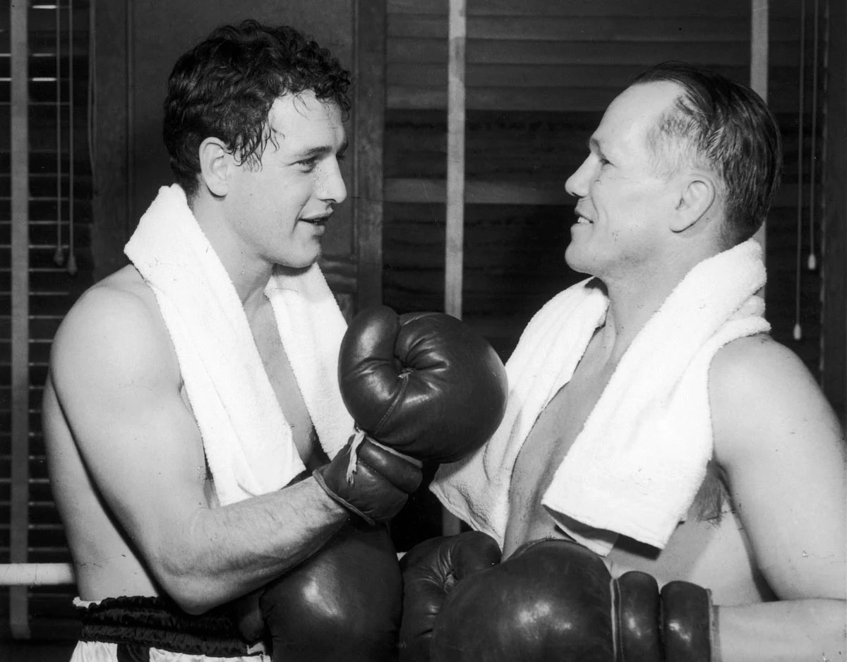 Paul Newman w Między liniami ringu Dwóch bokserów w rękawicach bokserskich rozmawia ze sobą twarzą w twarz, obaj z uśmiechami na twarzach i ręcznikami przewieszonymi przez szyje.