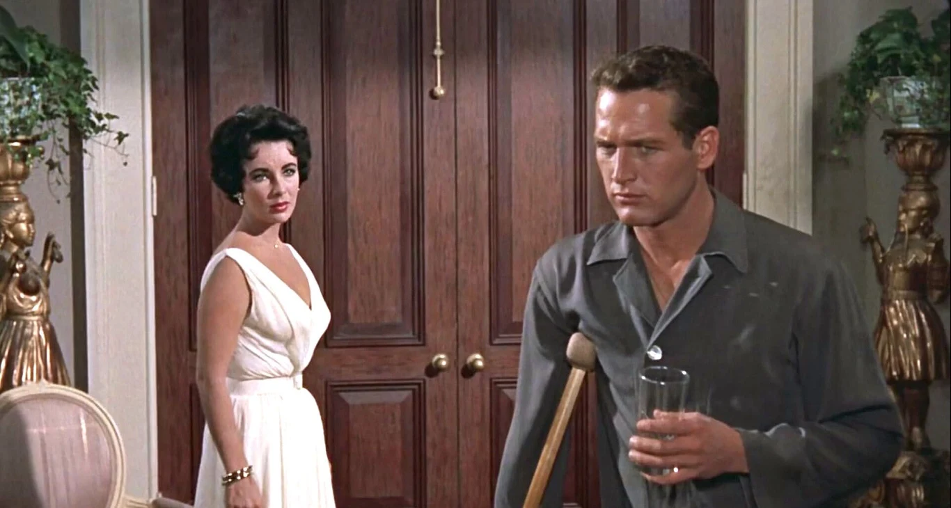 Liz Taylor i Paul Newman Mężczyzna o poważnym wyrazie twarzy z kulą pod pachą i szklanką w ręku stoi przed drewnianymi drzwiami, naprzeciwko niego kobieta w eleganckiej białej sukni spoglądająca z niepokojem.