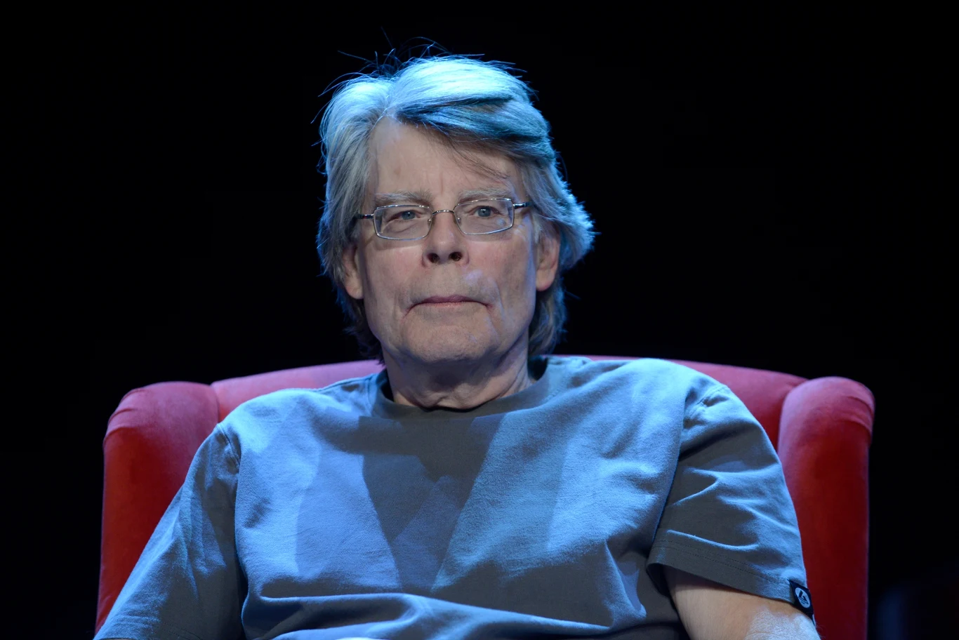 Stephen King Stephen King na czerwonym fotelu, w szarej koszulce, z siwymi włosami i okularami, na ciemnym tle