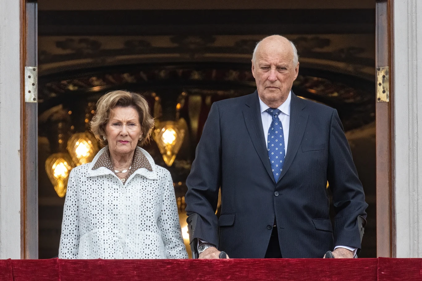 Starsza kobieta w jasnym płaszczu i mężczyzna w granatowym garniturze z niebieskim krawatem stoją obok siebie na balkonie, w tle widoczne ozdobne żyrandole.