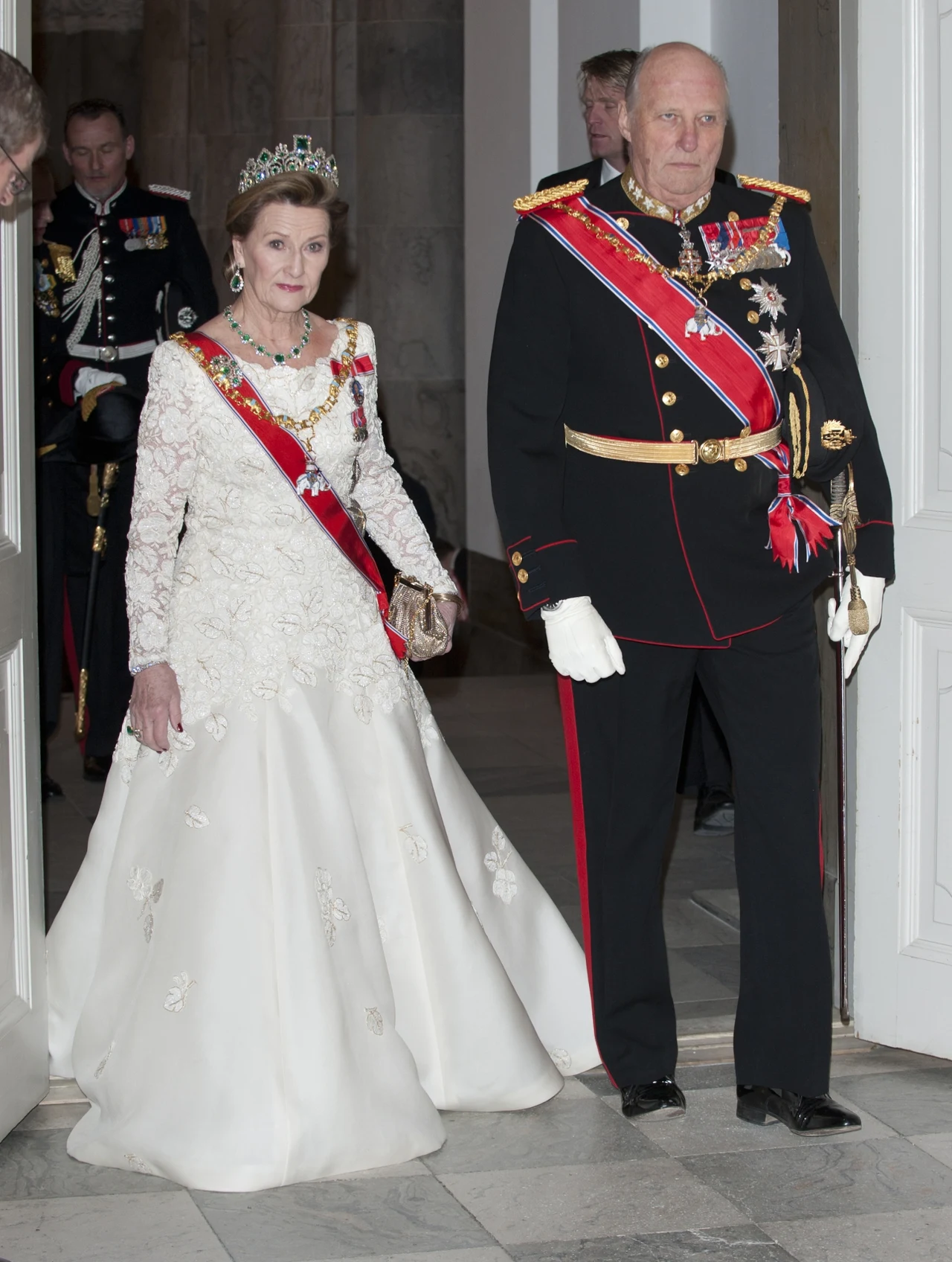 Para w strojach galowych: kobieta w białej sukni z koroną i ozdobami oraz mężczyzna w wojskowym mundurze z orderami i szarfą, oboje elegancko przygotowani na uroczyste wydarzenie.