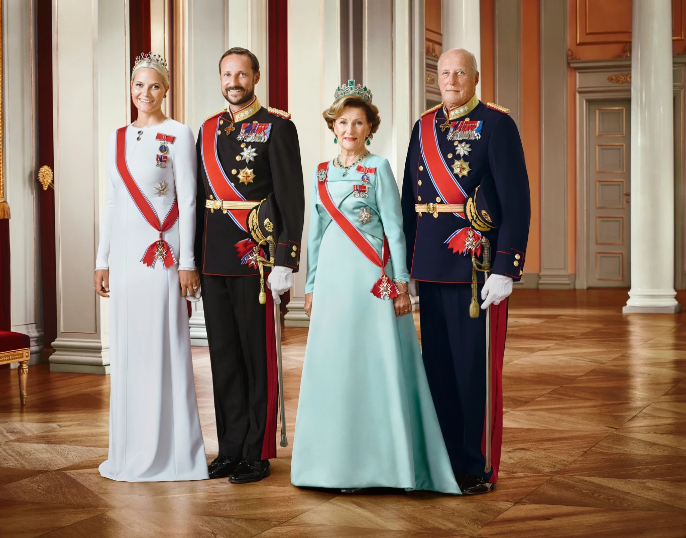 Cztery elegancko ubrane osoby w strojach galowych z odznaczeniami i tiarami pozują razem we wnętrzu pałacowym.
