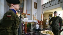 Dwóch żołnierzy w mundurach stoi po obu stronach udekorowanego katafalku w kościele, w otoczeniu świec ułożonych w kształt serca oraz kwiatów, na tle dużego krzyża i ołtarza.