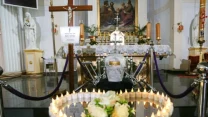 Trumna ze srebrną urną i krzyżem ustawiona przed ołtarzem, otoczona płonącymi świecami tworzącymi serce oraz bukietem białych róż, w tle ołtarz z obrazem religijnym.