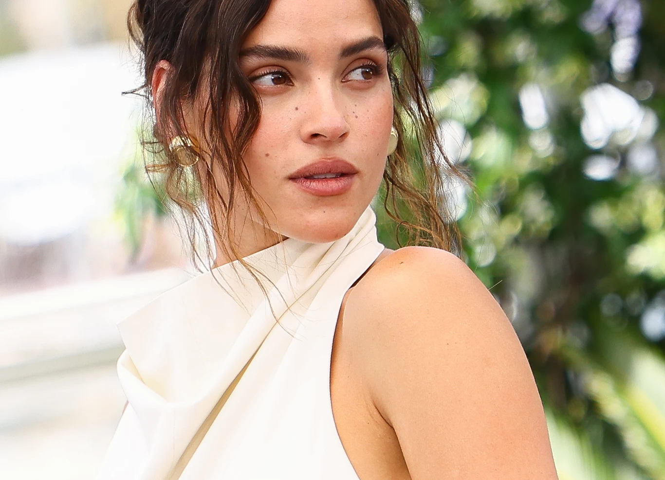 Adria Arjona Adria Arjona z ciemnymi, falowanymi włosami spiętymi luźno, w jasnej bluzce na jedno ramię, patrzy w bok.