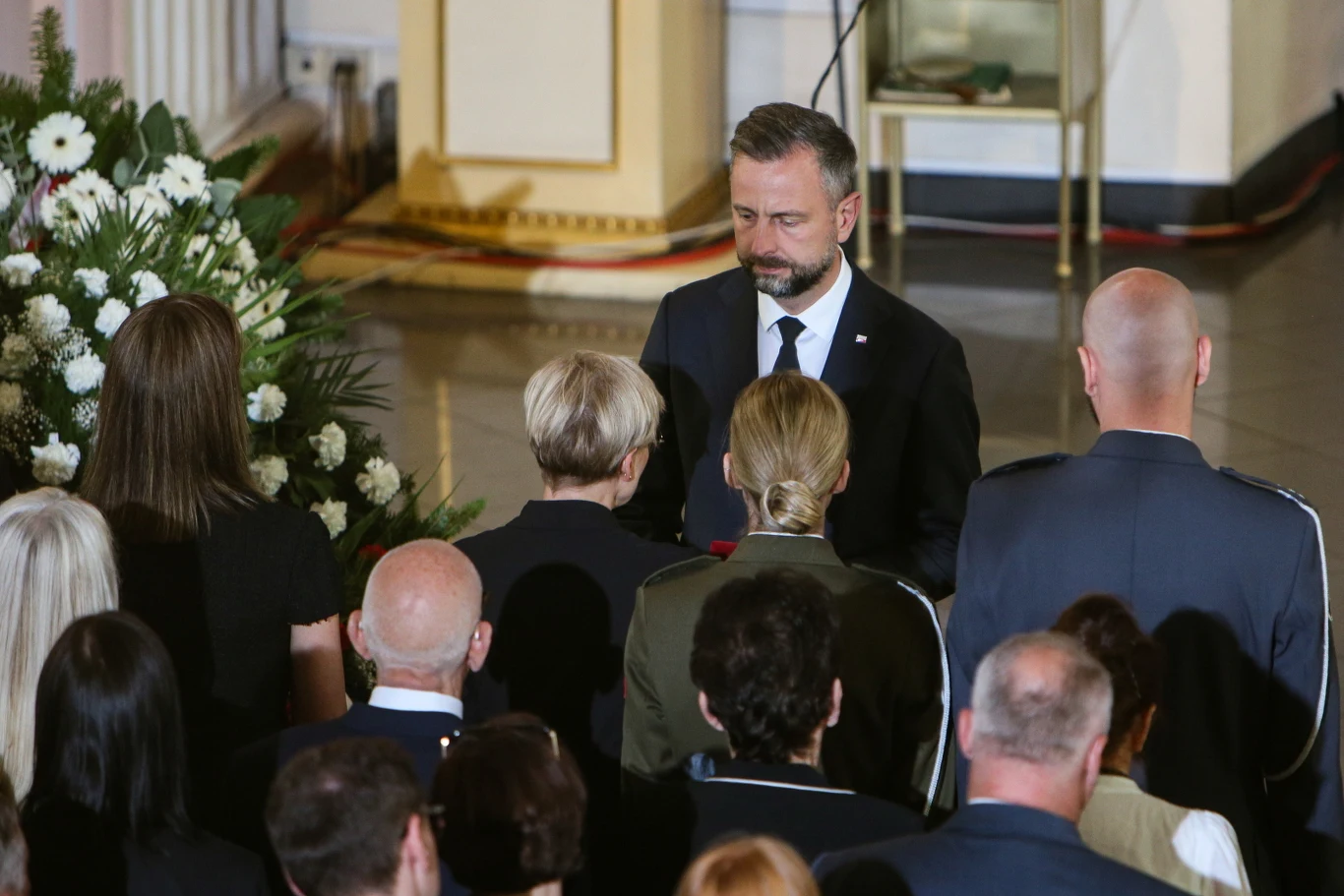 Władysław Kosiniak-Kamysz na pogrzebie gen. Mirosława Różańskiego Mężczyzna w garniturze stoi naprzeciw grupy ludzi w eleganckich ubraniach, otoczony wieńcami i kwiatami, w zabytkowym wnętrzu z wysokimi ścianami.