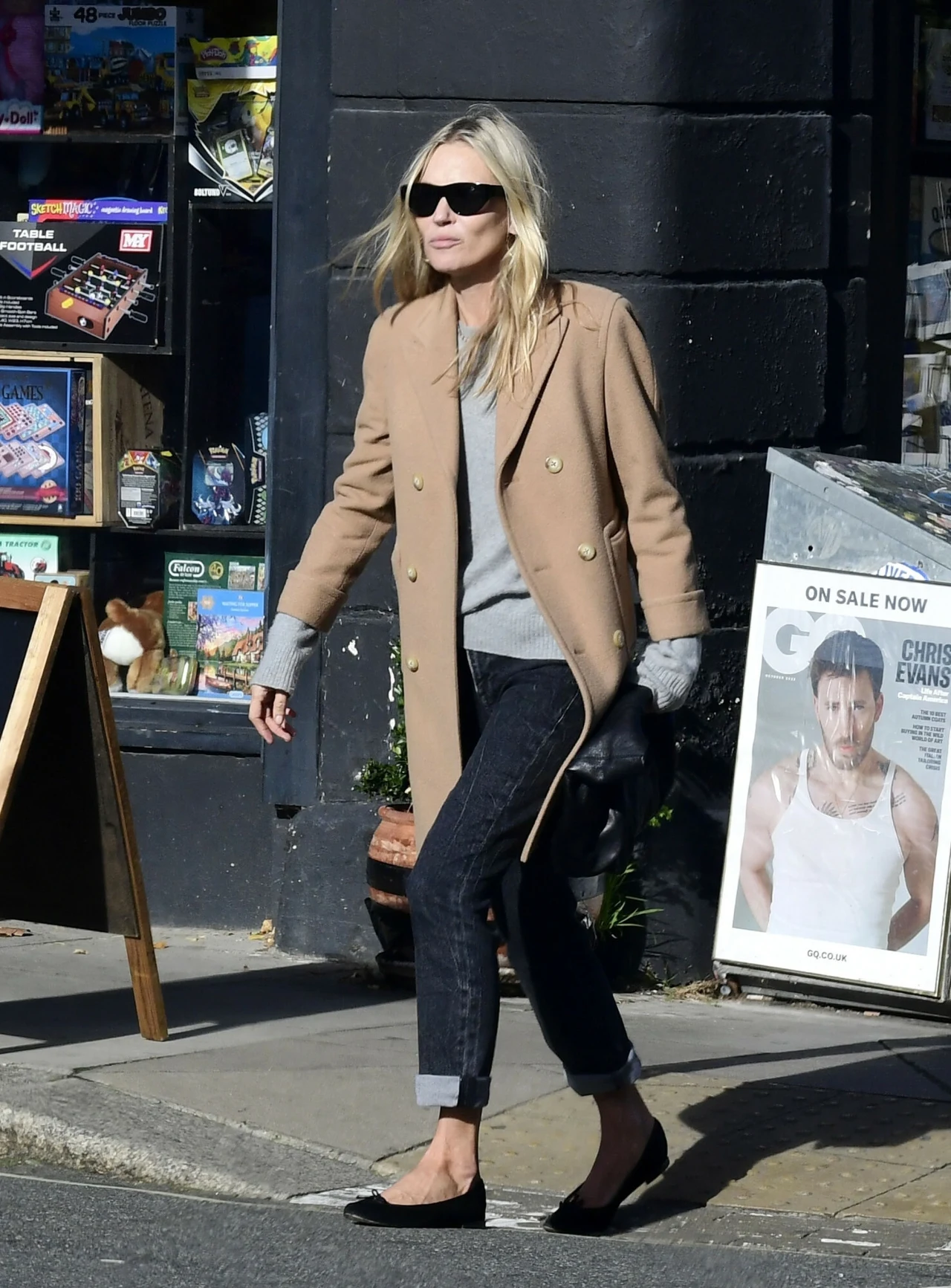 Kate Moss w beżowym płaszczyku Kobieta w beżowym płaszczu, szarym swetrze i czarnych okularach przeciwsłonecznych przechodzi chodnikiem obok kiosku z prasą; obok znajduje się plakat z magazynu GQ z wizerunkiem mężczyzny w białej koszulce.