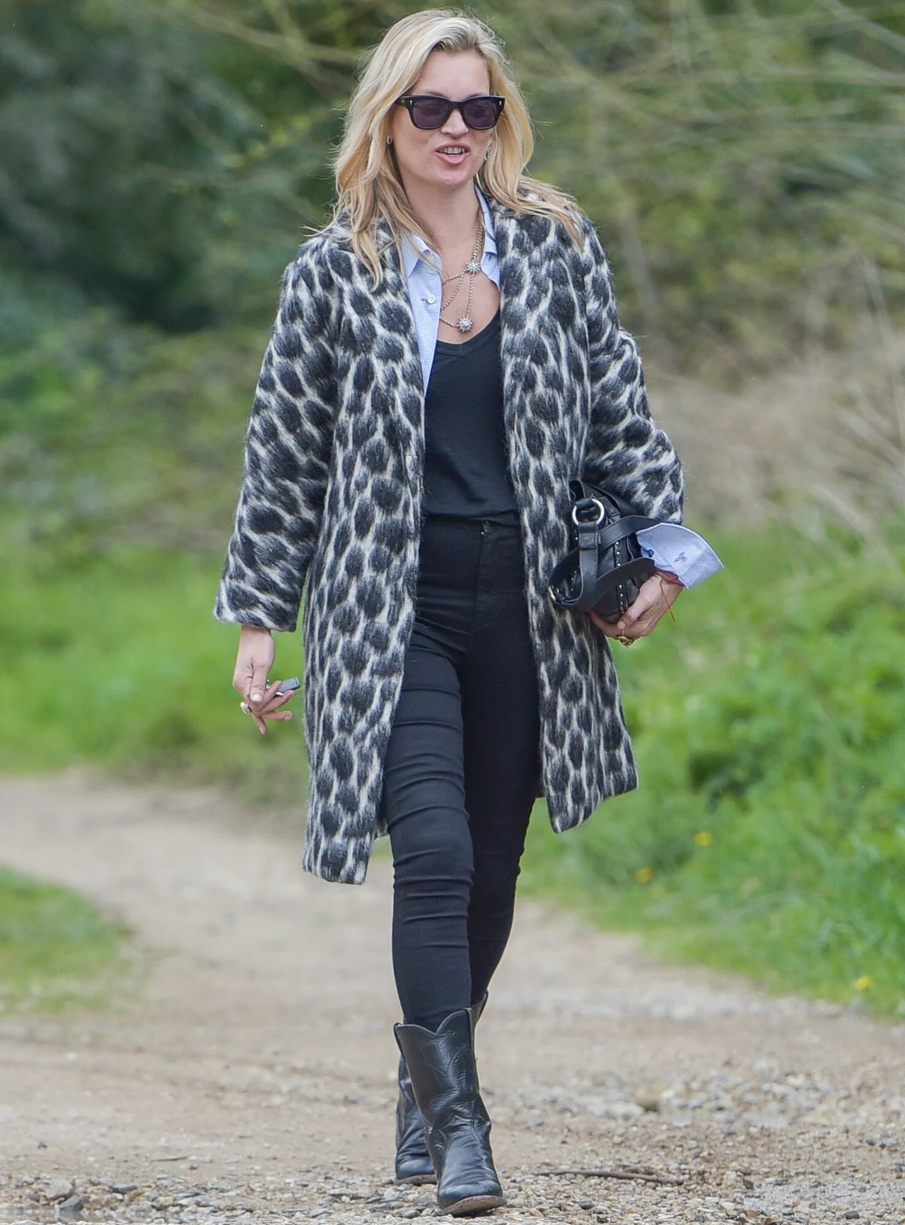 Kate Moss w płaszczu w cętki Kobieta w okularach przeciwsłonecznych, ubrana w wzorzysty płaszcz, czarne spodnie i buty, z torebką przewieszoną na ramieniu, spaceruje wiejską ścieżką.