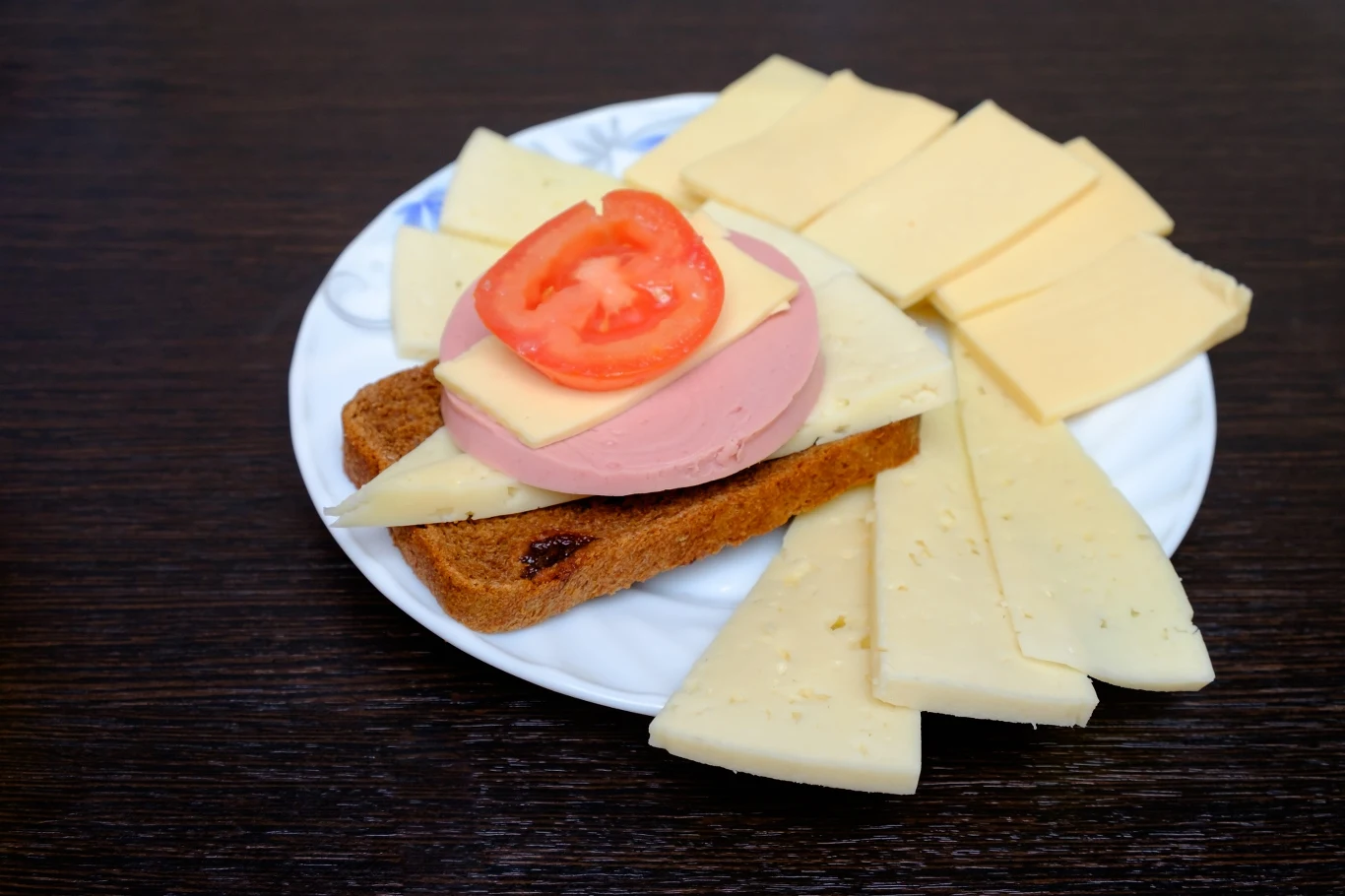 Warto przede wszystkim wybierać nabiał wysokiej jakości, z jak najkrótszym składem i łączyć go z innymi, wartościowymi produktami Tost z jasnego pieczywa z plasterkiem kiełbasy, żółtym serem i plastrem pomidora, otoczony kawałkami sera żółtego na białym talerzu.