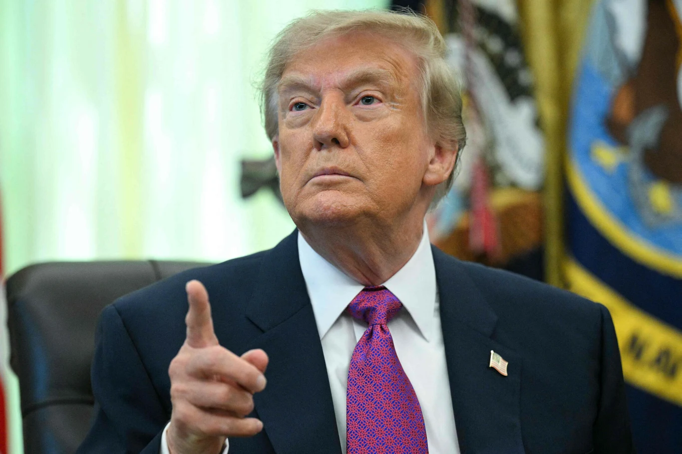 Co, jeśli geograficzna kreatywność prezydenta USA będzie nadal rosła? Mężczyzna w garniturze i czerwonym krawacie siedzi przy biurku, skierowany lekko w bok, z uniesionym palcem wskazującym, w tle widoczne flagi i emblematy.