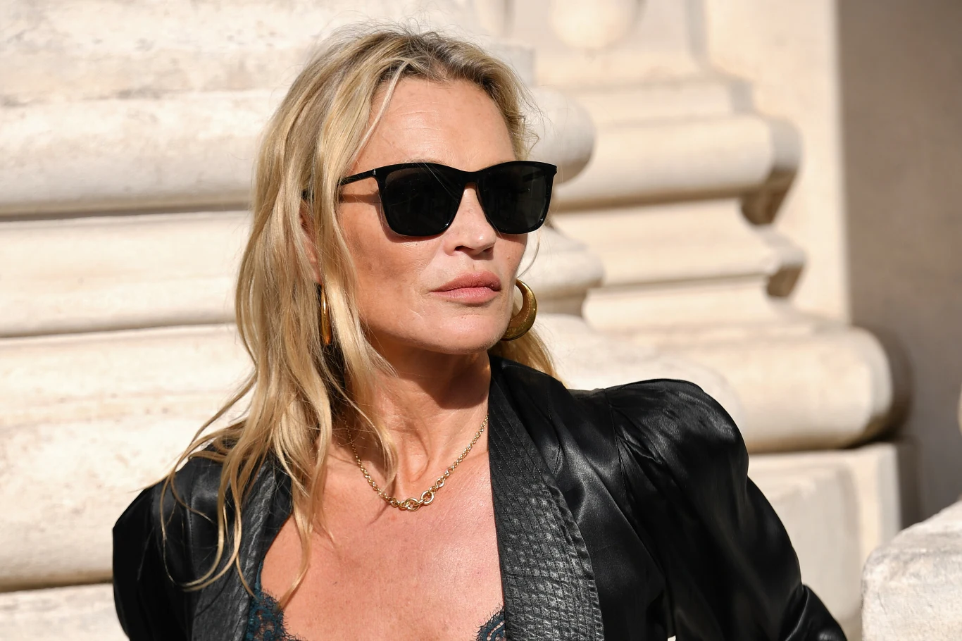 Kate Moss to wciąż jedna z najlepszych modelek na świecie Kate Moss w czarnych okularach przeciwsłonecznych, czarnej satynowej marynarce i złotej biżuterii na tle jasnej architektury.