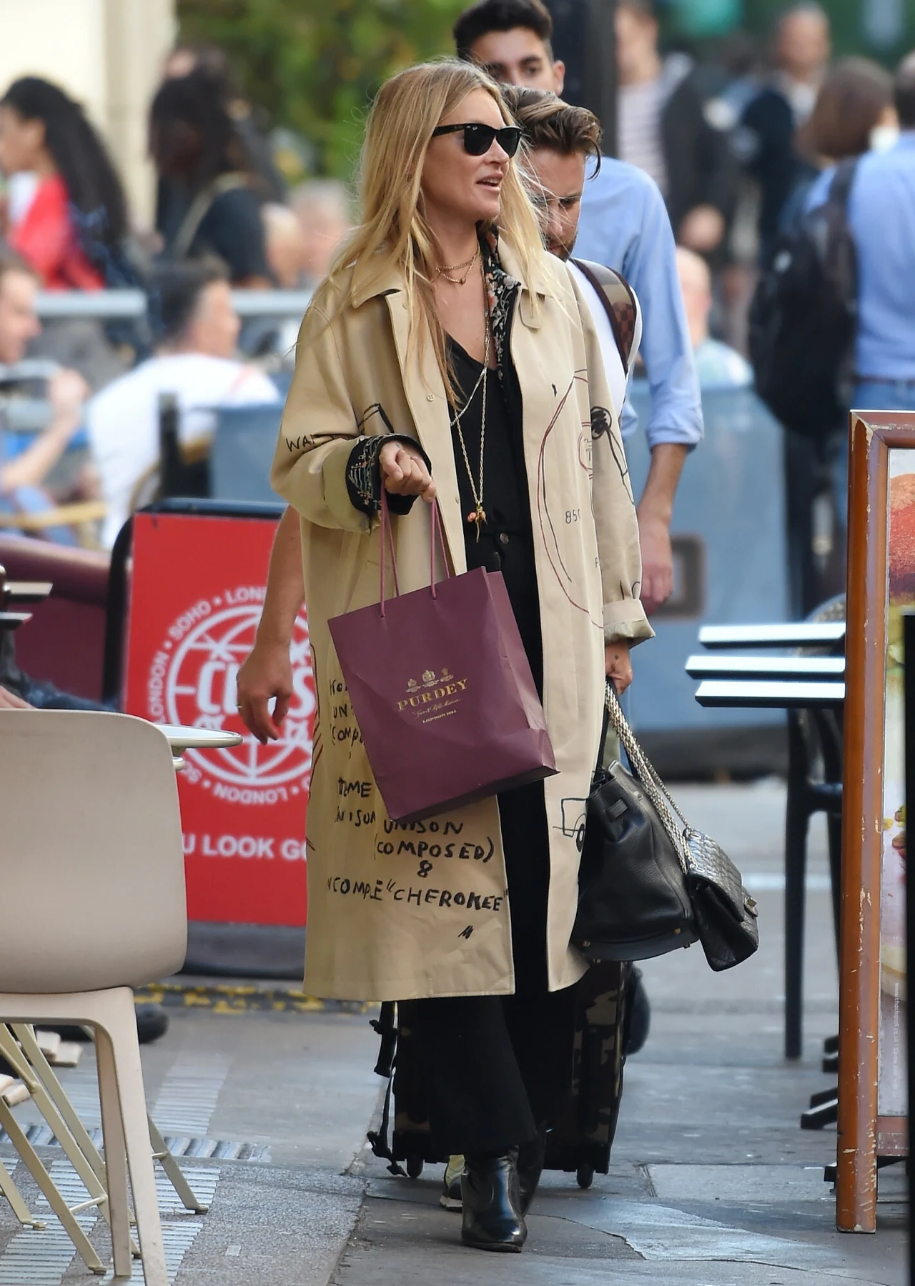 Kate Moss w beżowym, stylowym płaszczu Kobieta w jasnym płaszczu, ciemnych okularach i długiej sukience niesie papierową torbę zakupową oraz czarną torebkę, idąc po chodniku miejskiej ulicy wśród przechodniów.