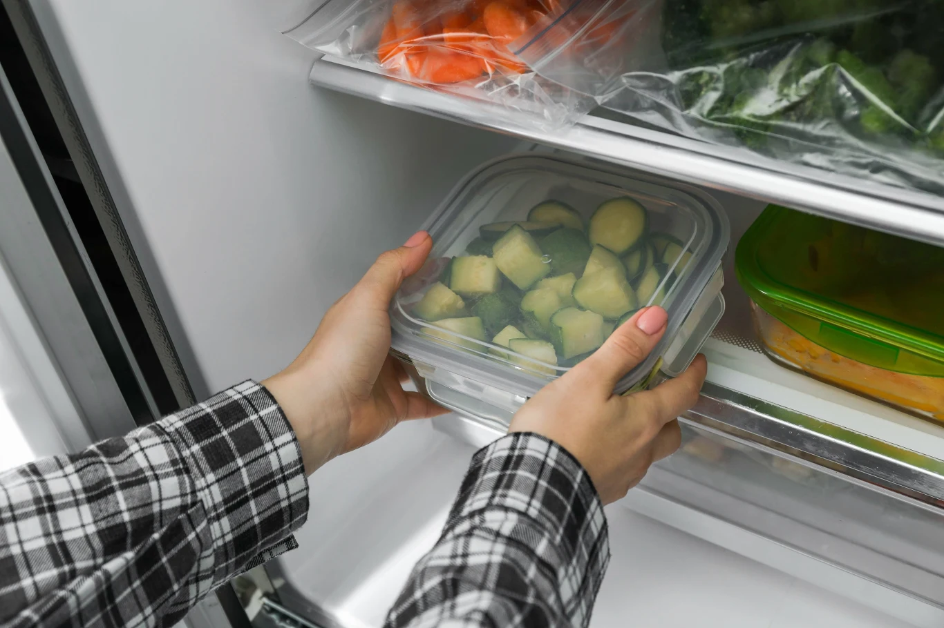 Ręce wkładają plastikowy pojemnik z pokrojonymi warzywami na półkę w lodówce, obok innych zamkniętych pojemników spożywczych.