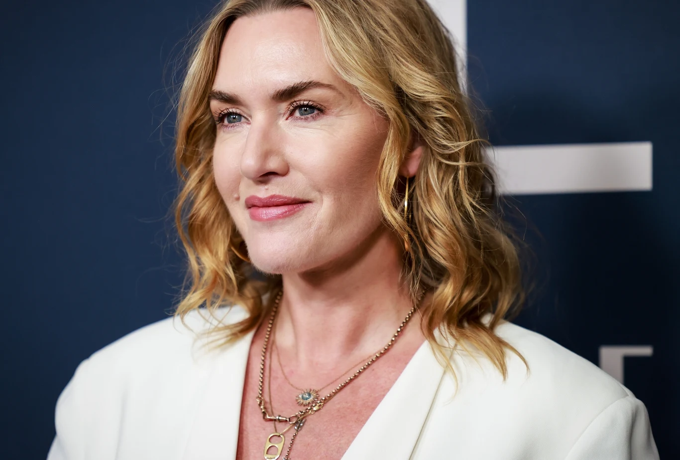 Kate Winslet Kate Winslet w białej marynarce i złotych naszyjnikach na tle granatowej ścianki z geometrycznymi wzorami