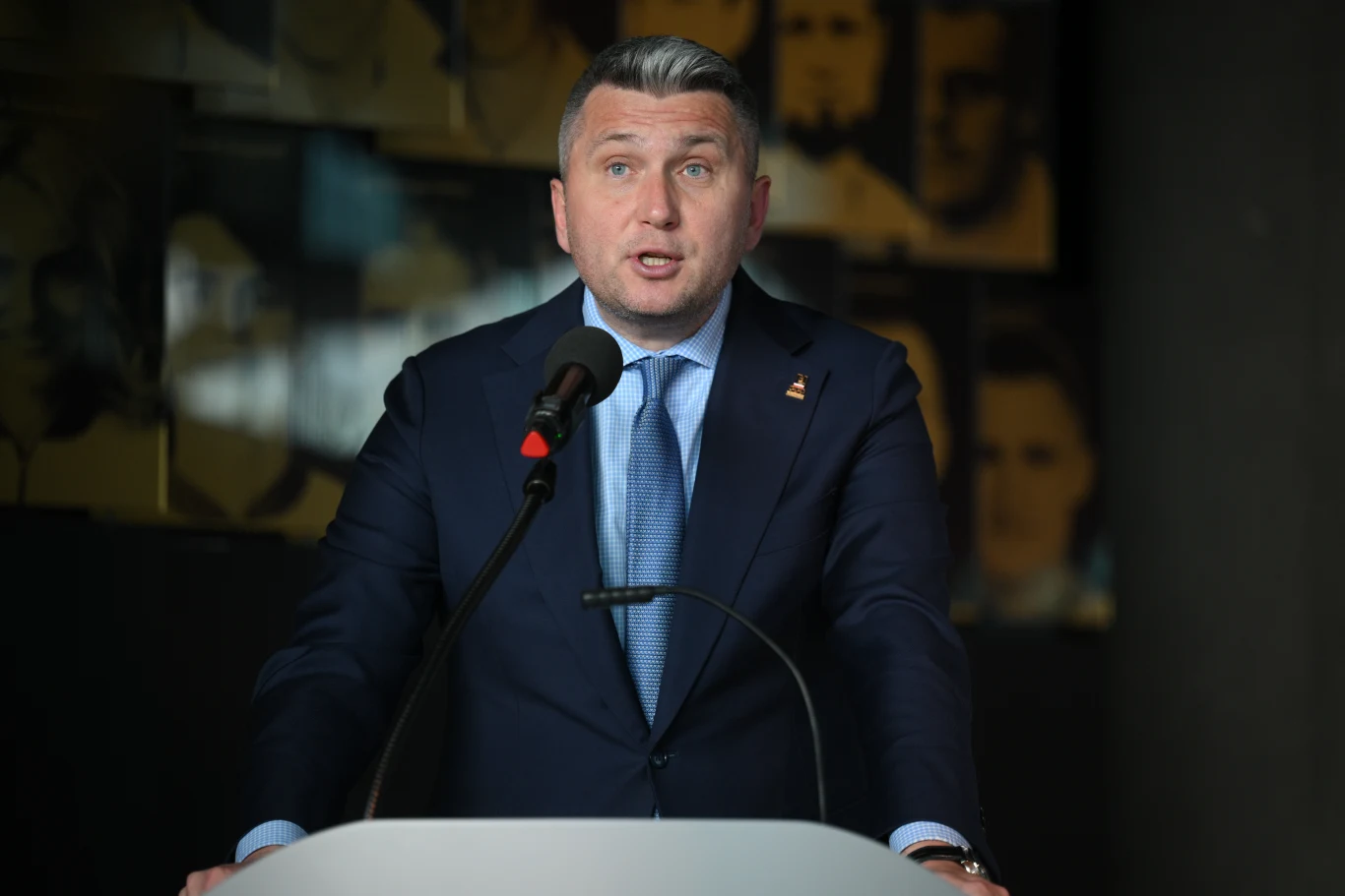 Prezes PKOl Radosław Piesiewicz Prezes PKOl Radosław Piesiewicz w garniturze i niebieskim krawacie przemawia za mównicą na tle portretów