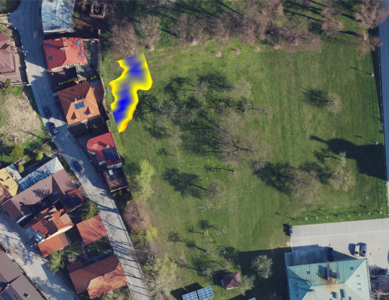 Duży trawnik otoczony drzewami oraz domami, z wyraźnie zaznaczoną żółto-fioletową plamą w pobliżu zabudowań przy lewej krawędzi zdjęcia.