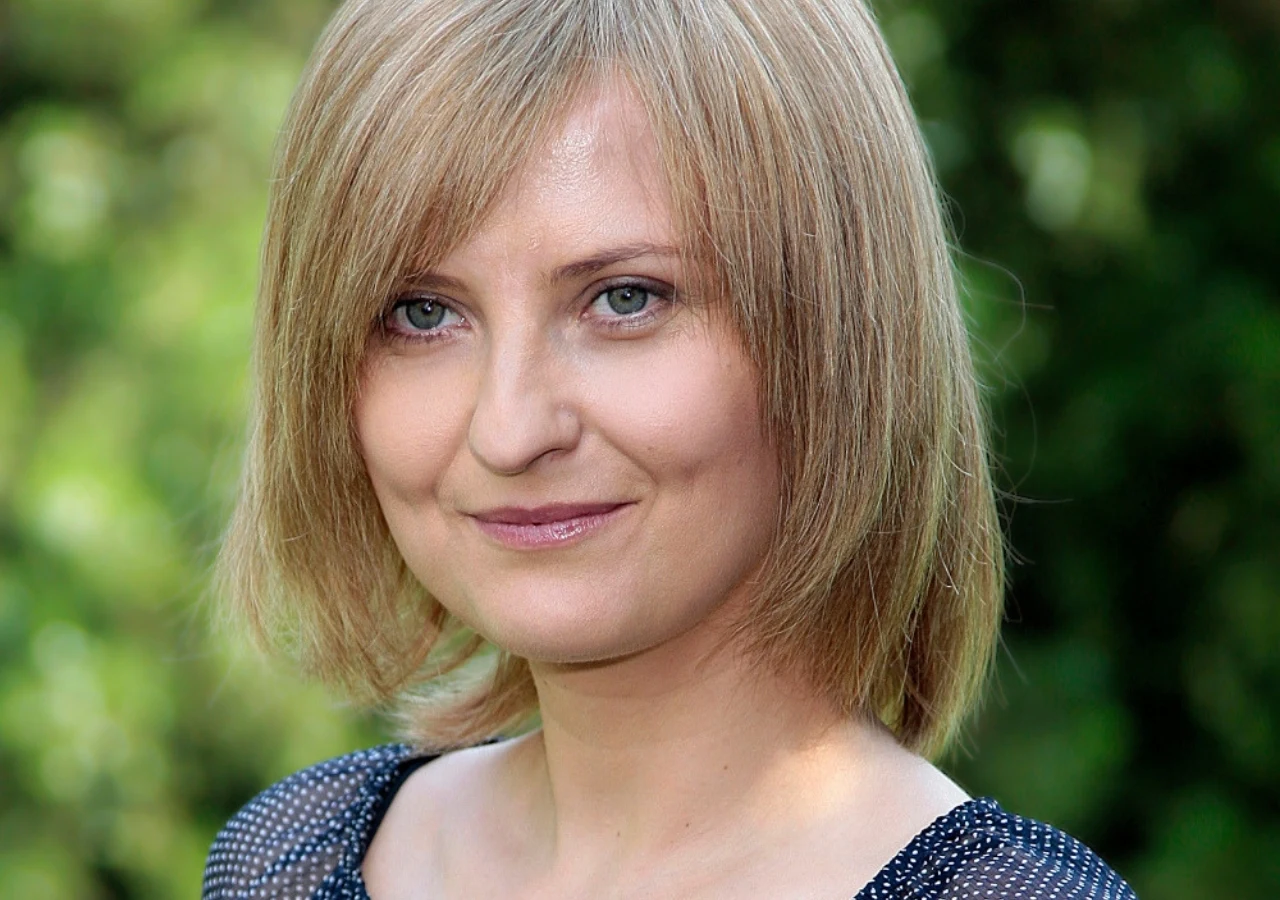 Kobieta o jasnych, prostych włosach do ramion i niebieskich oczach, lekko uśmiechnięta, ubrana w bluzkę z drobnym wzorem, sfotografowana w naturalnym świetle na tle zieleni.