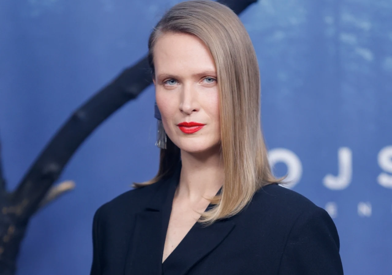 Kobieta o blond włosach w eleganckiej, ciemnej marynarce, z wyraźnie zaznaczonymi czerwonymi ustami, patrząca prosto w obiektyw na tle ciemnoniebieskiej scenerii.