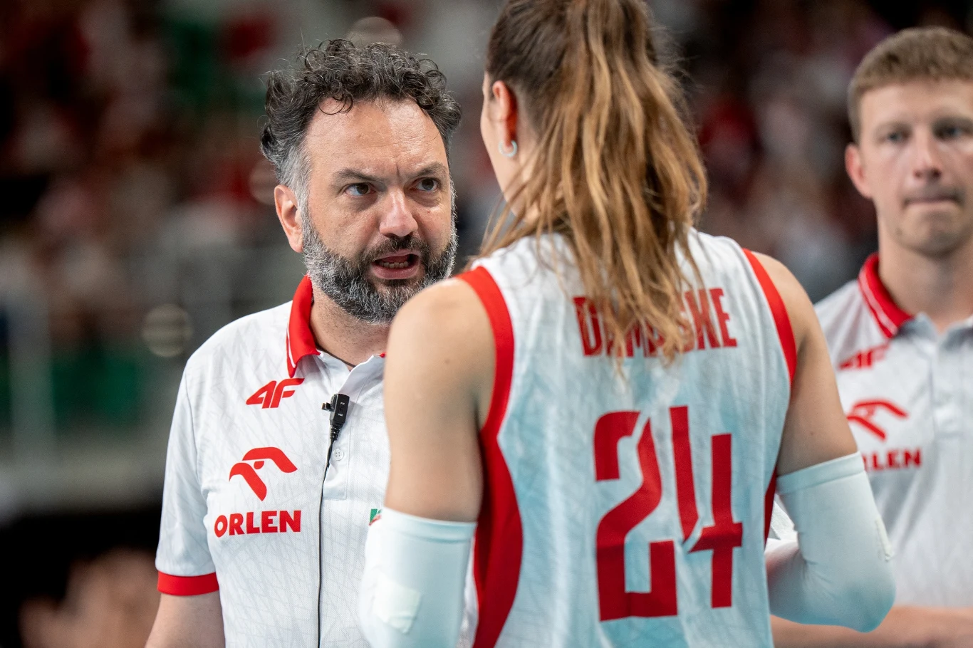 Trener drużyny siatkarek w białym stroju sportowym rozmawia z zawodniczką mającą na koszulce numer 24 oraz charakterystyczny napis, obok stoi kolejny członek zespołu w podobnym stroju.