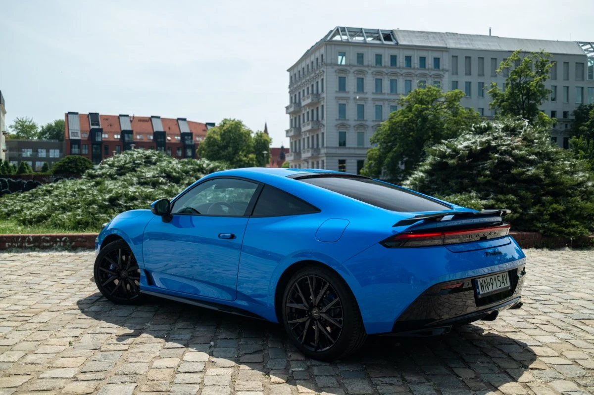 Wysiadając na ciasnym parkingu trzeba uważać na długie drzwi. Niebieskie sportowe coupe z przyciemnianymi szybami i czarnymi felgami zaparkowane na brukowanym placu w otoczeniu zieleni oraz miejskich budynków.