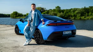 Mężczyzna w jasnym ubraniu opiera się o niebieski, sportowy samochód zaparkowany nad jeziorem, w otoczeniu zielonych drzew i nieba bez chmur.