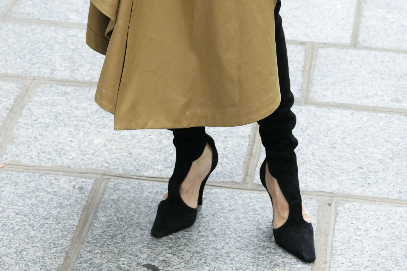 Celine Dion w zamszowych kozakach. Oryginalne buty przyciągają wzrok Czarne, zamszowe kozaki na wysokim obcasie o nietypowym wycięciu, zestawione z długim, beżowym płaszczem na chodniku z szarych płyt.