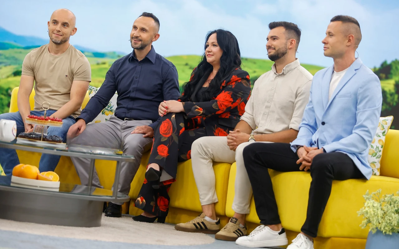 Pięć osób siedzących na żółtej kanapie w studiu telewizyjnym, z przodu szklany stolik z talerzem ciasta i miską owoców.