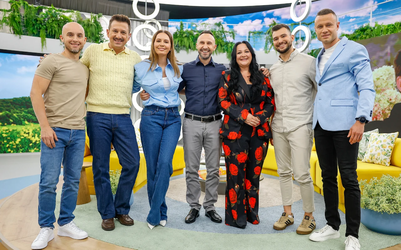 Siedmioro uśmiechniętych osób stoi razem w jasnym, nowoczesnym studio telewizyjnym na tle zieleni i dużego ekranu z widokiem na naturę; w centrum znajduje się kobieta w czarnym kombinezonie w czerwone kwiaty, obok niej mężczyźni i kobieta w casualowych...