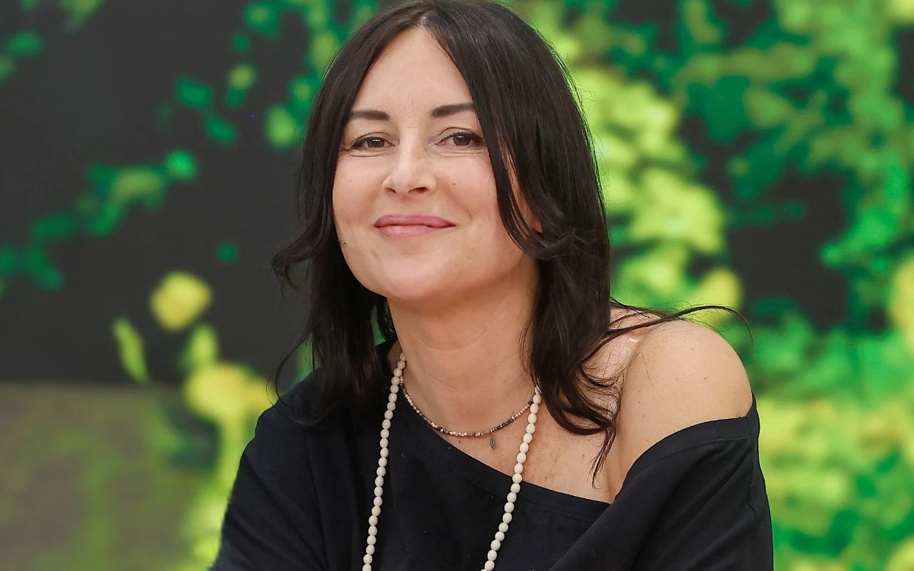 Emilia Kostrzewska Kobieta o ciemnych włosach, ubrana w czarną bluzkę z odsłoniętym jednym ramieniem i naszyjnik z białych koralików.