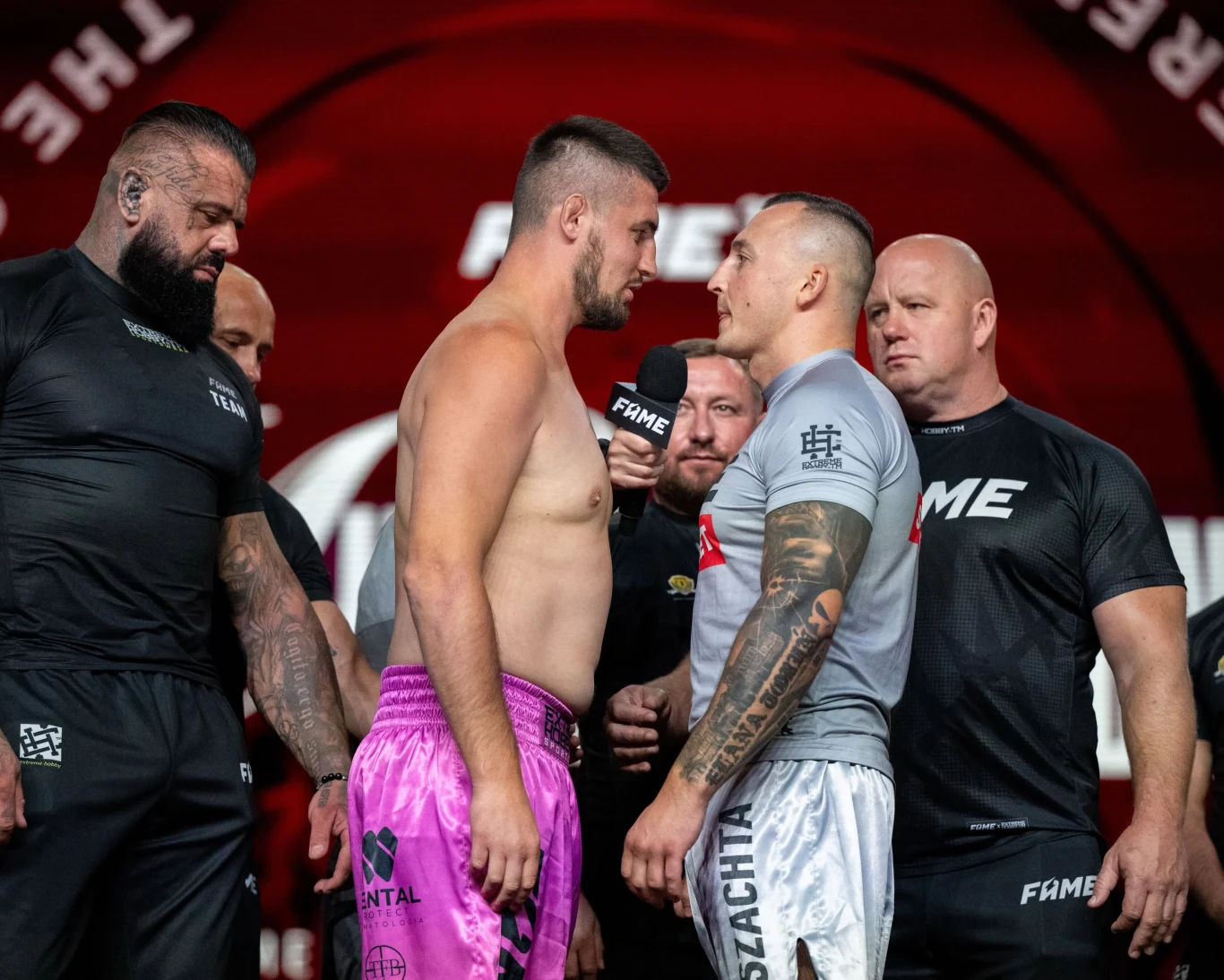 FAME MMA 32: Wyniki NA ŻYWO. Kasjusz Don Kasjo Życiński - Bartosz Szachta Dwóch zawodników stojących naprzeciwko siebie twarzą w twarz podczas ważenia, otoczonych przez ochroniarzy i organizatorów wydarzenia sportowego.
