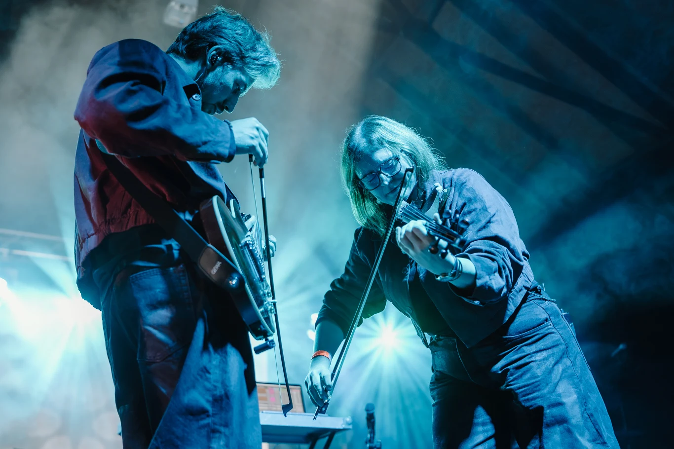 Imasleep Dwoje muzyków grających na scenie, jeden na gitarze elektrycznej, drugi na skrzypcach w akompaniamencie efektownego oświetlenia scenicznego.