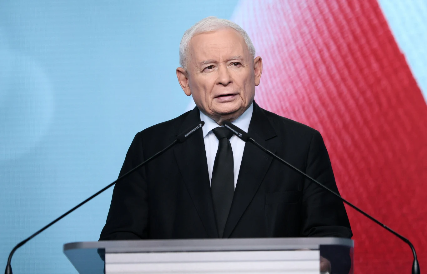 Jarosław Kaczyński Jarosław Kaczyński przy mównicy z dwoma mikrofonami na tle biało-czerwonej flagi.