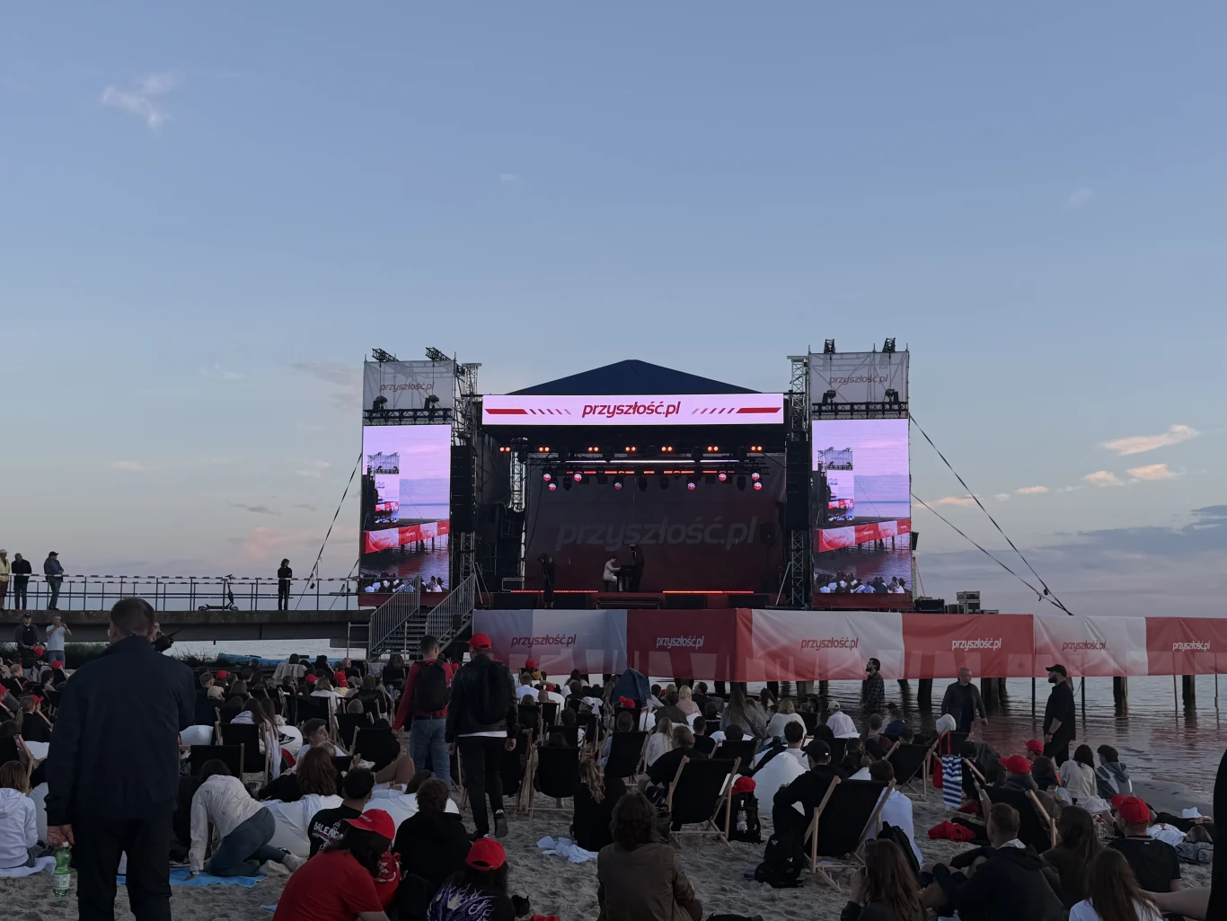Scena z dużymi ekranami LED ustawiona na plaży przy brzegu morza, otoczona przez tłum siedzących i stojących osób uczestniczących w wydarzeniu.