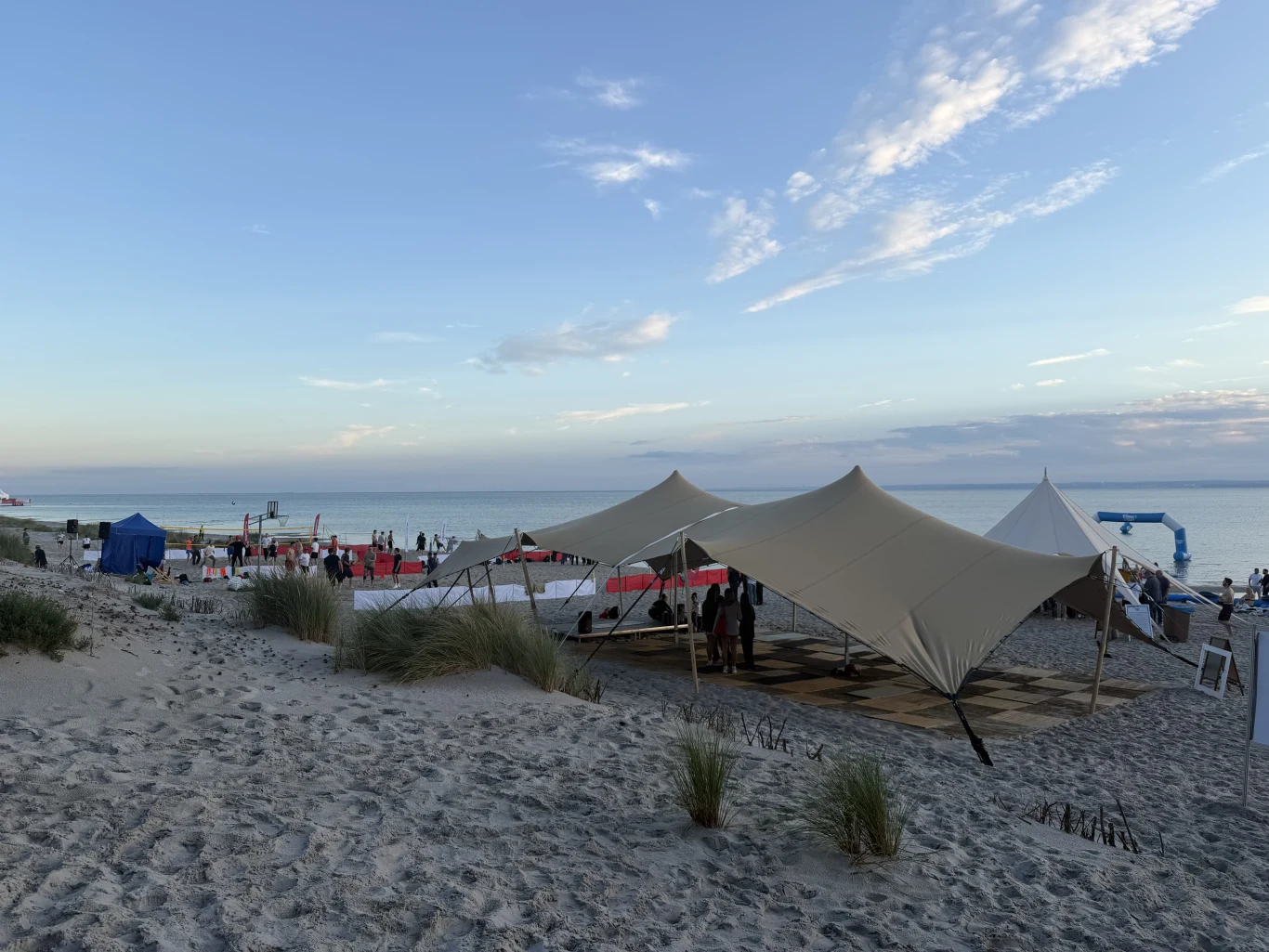 Plaża z dużym namiotem rozstawionym na piasku, grupą ludzi pod zadaszeniem i jasnym niebem nad spokojnym morzem.