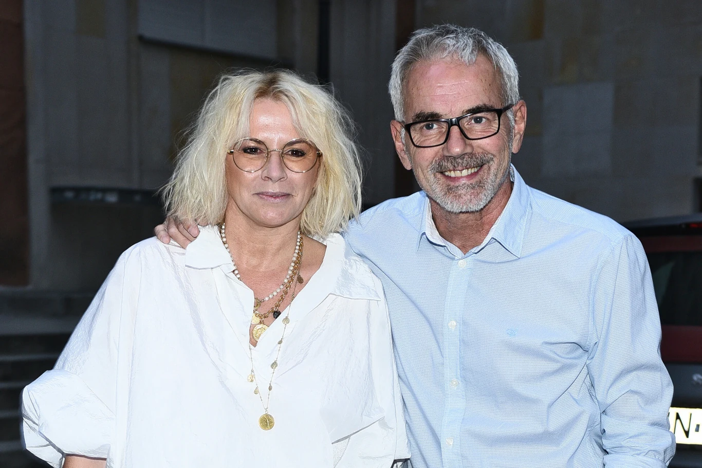 Robert Janowski i jego żona Monika Dwie uśmiechnięte osoby stojące blisko siebie, obie w jasnych koszulach; kobieta z blond włosami i okrągłymi okularami, mężczyzna z siwymi włosami i brodą, w okularach, obejmują się przyjacielsko.