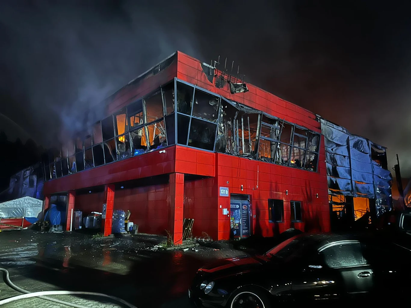 Pożar hali w Gdyni Dwupiętrowy budynek o czerwonej elewacji i dużych przeszklonych oknach, znacznie zniszczony przez pożar, z widocznymi zwęglonymi fragmentami ścian i wewnętrznym światłem ognia.