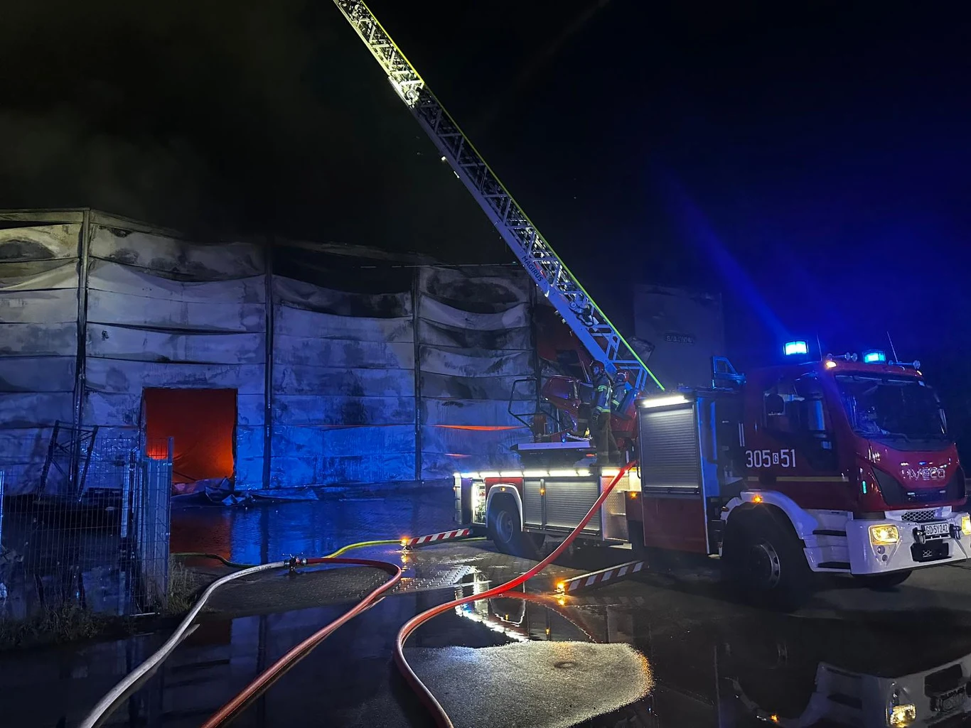 Pożar hali w Gdyni Wielki pożar hali przemysłowej, straż pożarna prowadzi akcję gaśniczą z użyciem wozu strażackiego i drabiny mechanicznej, wokół rozłożone są węże gaśnicze.