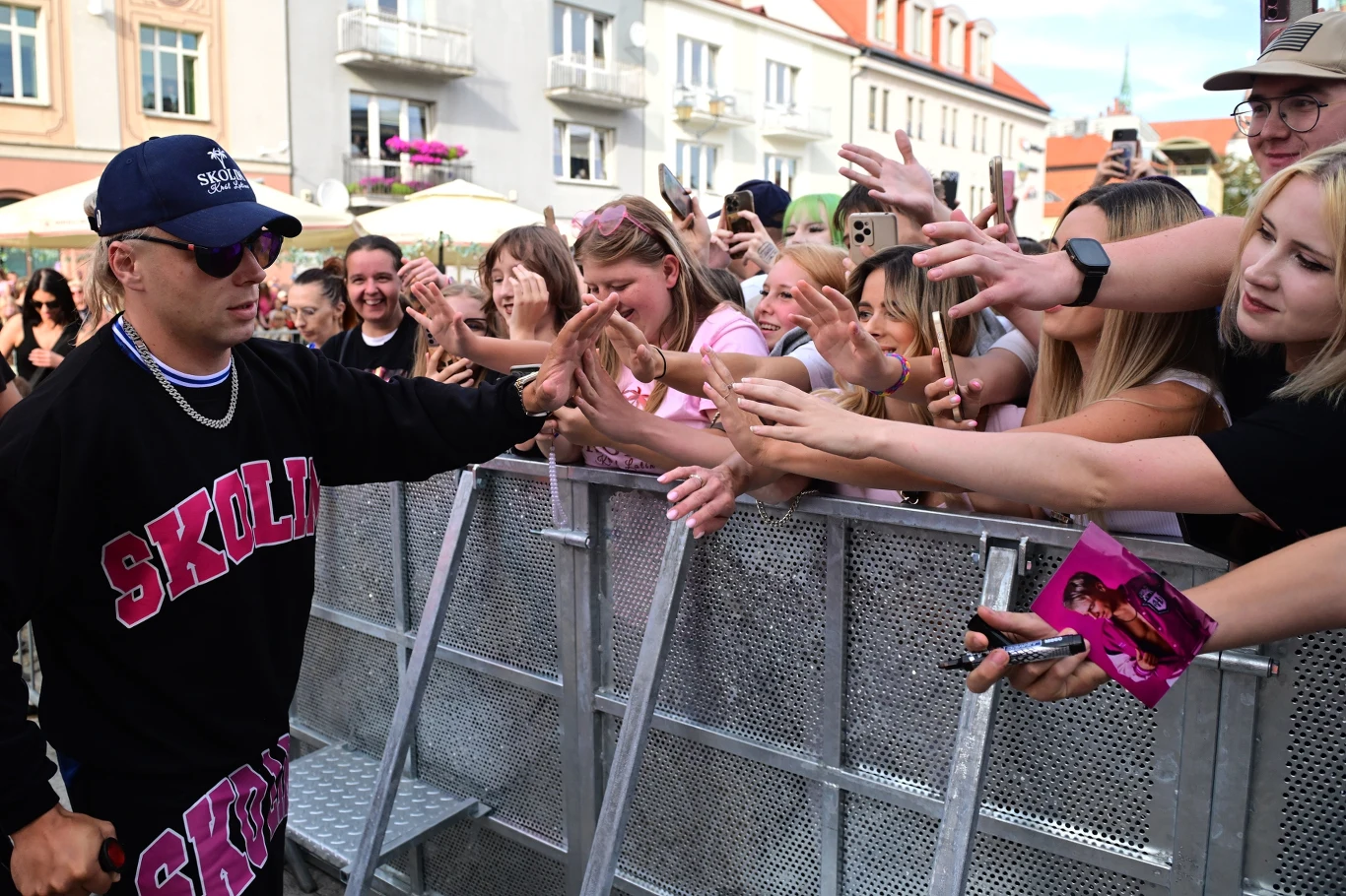 Skolim i jego młodzi fani Skolim w czapce i okularach podaje ręce fanom przy barierkach, tłum wyciąga dłonie w jego stronę.