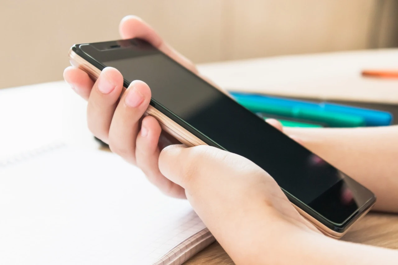Dłoń trzymająca smartfon nad otwartym zeszytem, w tle leżą kolorowe długopisy.