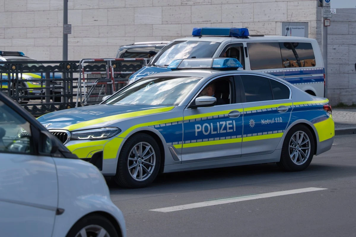 Niemieckie urzędy mają obecnie sześć miesięcy na ściganie większości wykroczeń drogowych. Nowy termin dotyczy również kierowców z Polski. Radiowóz niemieckiej policji z oznaczeniami i sygnałami świetlnymi patroluje miasto, w tle policyjny van.