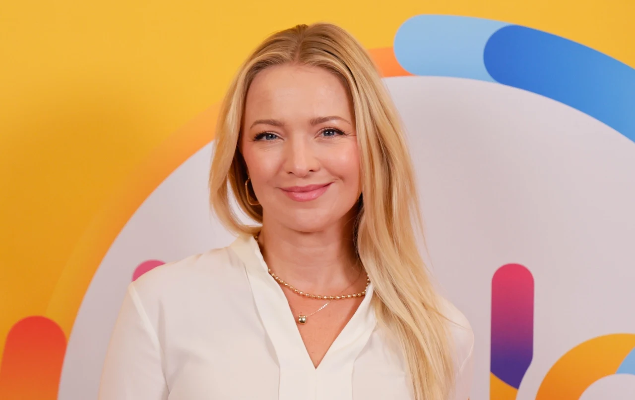 Barbara Kurdej-Szatan w "halo tu polsat" Barbara Kurdej-Szatan w białej bluzce, długie blond włosy, na tle żółtego plakatu z kolorowymi okręgami i liniami.
