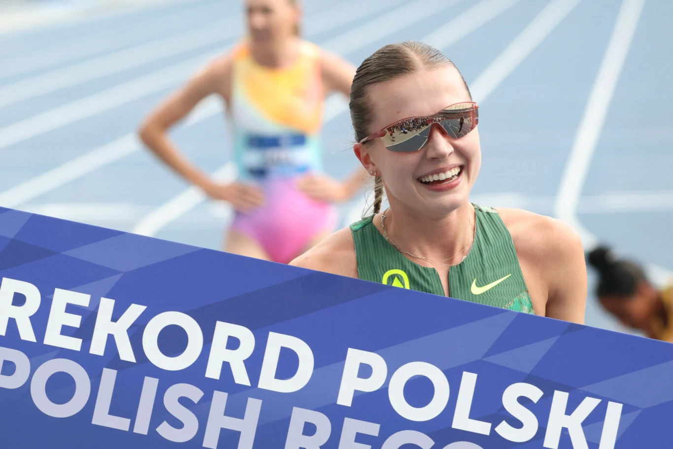 Klaudia Kazimierska tydzień temu poprawiła rekord Polski na 1500 metrów. Dziś atakowała ten na 800 metrów Uśmiechnięta lekkoatletka w zielonym stroju sportowym i okularach przeciwsłonecznych, trzymająca transparent z napisem 'Rekord Polski'.