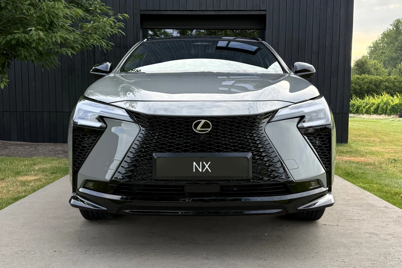 Lexus NX po liftingu został wyposażony m.in. w nową, bezramkową osłonę chłodnicy. Szara Toyota Lexus NX zaparkowana przed nowoczesnym budynkiem, widok od frontu podkreślający duży czarny grill i charakterystyczne przednie reflektory LED.