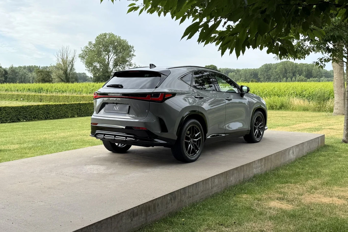 Pierwsze egzemplarze Lexusa NX zaczną spływać do Europy wiosną 2027 r. Szary SUV zaparkowany na betonowym podwyższeniu, otoczony zielonym trawnikiem i polami w tle.